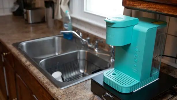 Una cocina con una cafetera color turquesa sobre una base negra, un lavamanos de acero inoxidable y una ventana con cortinas.