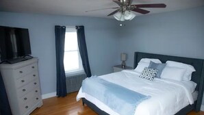 Un dormitorio con cama, cómoda, televisor, ventilador de techo y ventana con cortinas.