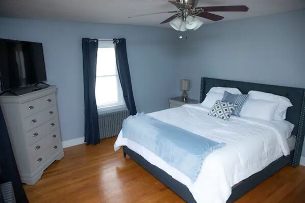 A bedroom with a bed, a dresser, a TV, a ceiling fan, and a window with curtains.