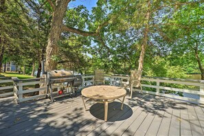 Une terrasse avec une table ronde et des chaises, un barbecue et une vue sur un lac entouré d’arbres.
