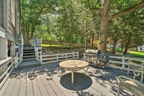 Une terrasse en bois avec une table ronde et des bancs, entourée d’arbres et agrémentée d’un barbecue.