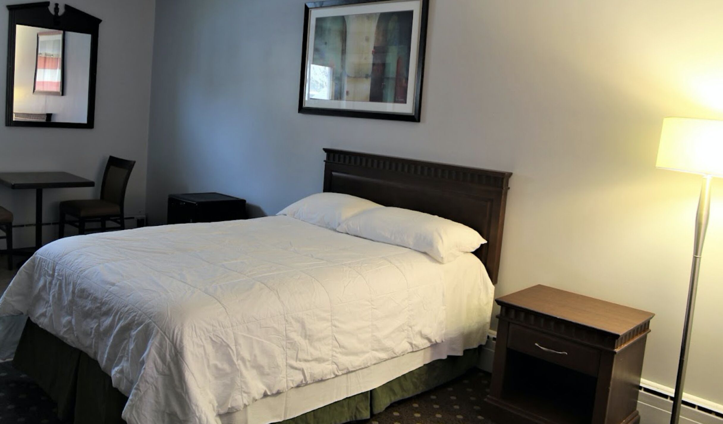 A hotel room with a large bed, a desk, a chair, a nightstand, and a lamp.