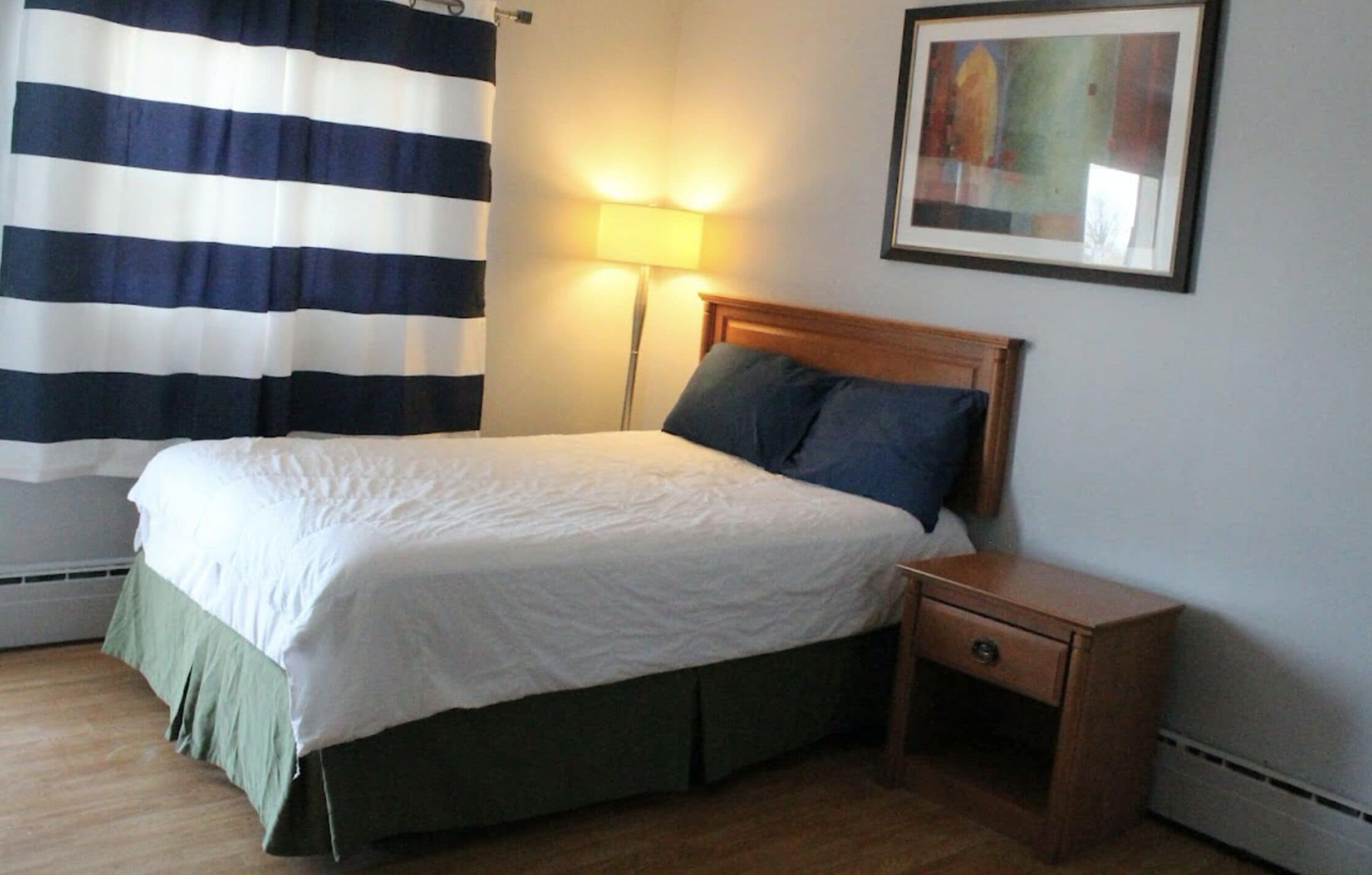 A neatly made bed with a wooden headboard, a bedside table with a drawer, a floor lamp, and a striped curtain.