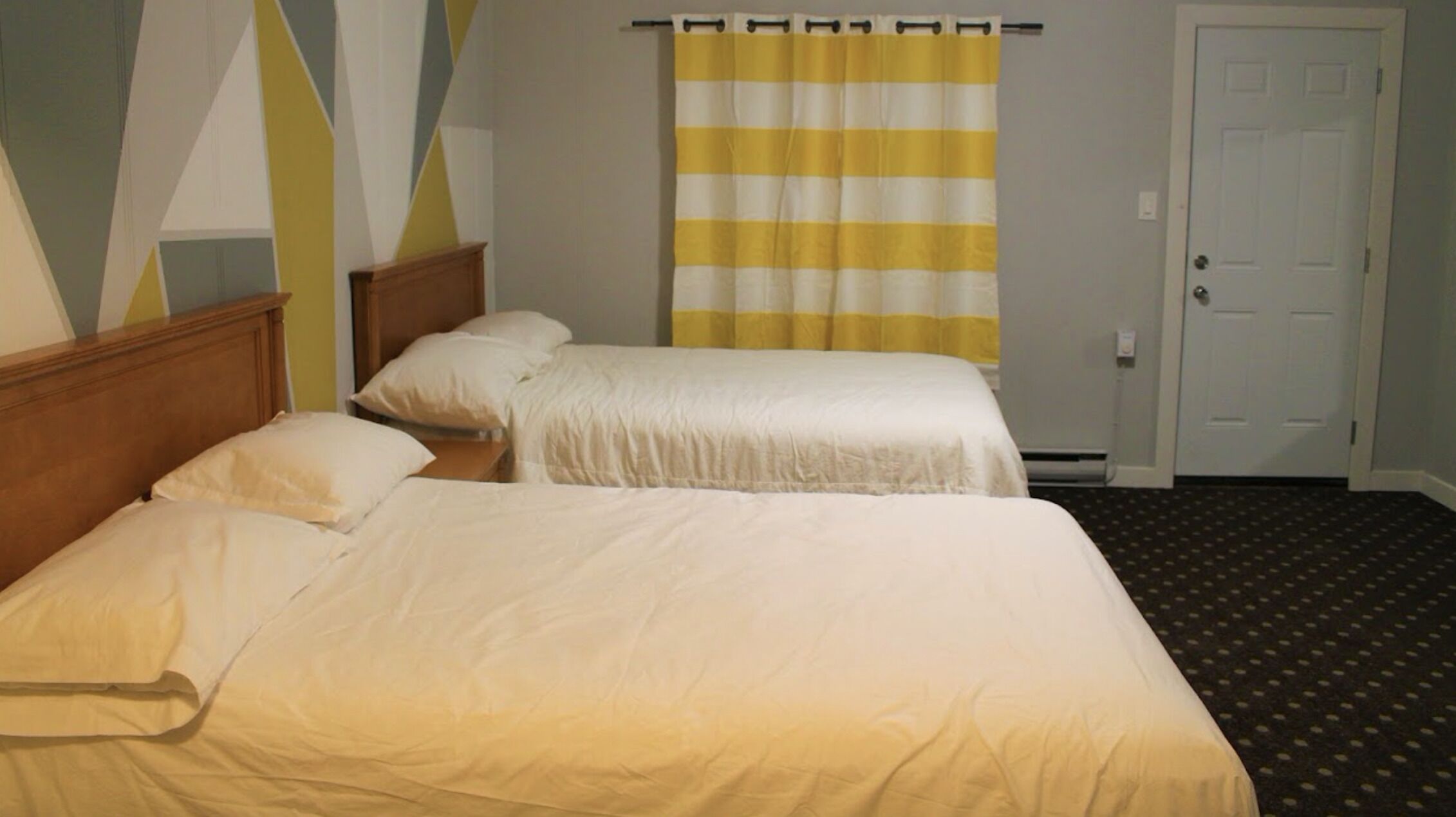 Habitación de hotel con dos camas, cabecera de madera, cortina a rayas amarillas y blancas, y una puerta con pomo.