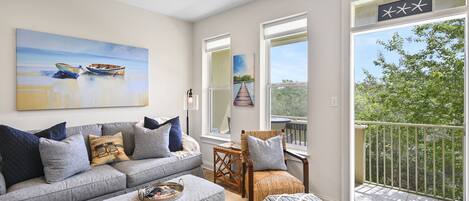 A modern living room with a grey sofa, ottoman, and wicker chair, a painting of boats, and a balcony with a railing.