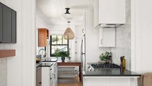 A modern kitchen with white cabinetry, a central island with stools, a stove, and a hanging light fixture. - Historic Beach Cottage with Ocean View Deck - Steps to Beach (Dillon Beach)