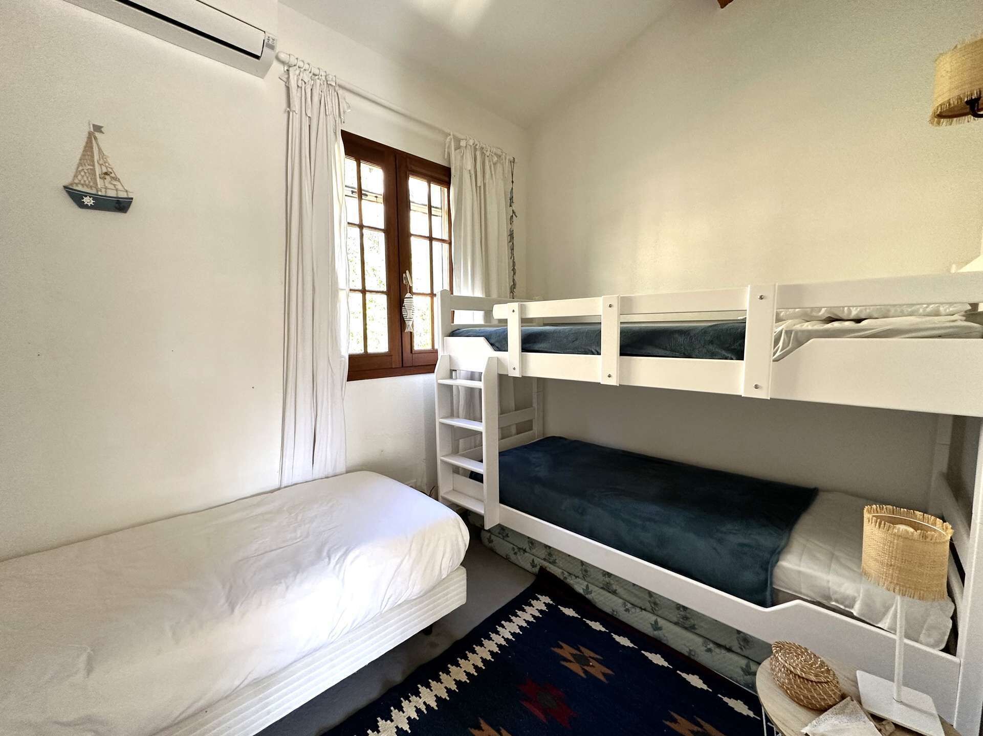 A room with a bunk bed, a single bed, a window with curtains, a small sailboat decoration, and a woven basket on a nightstand.