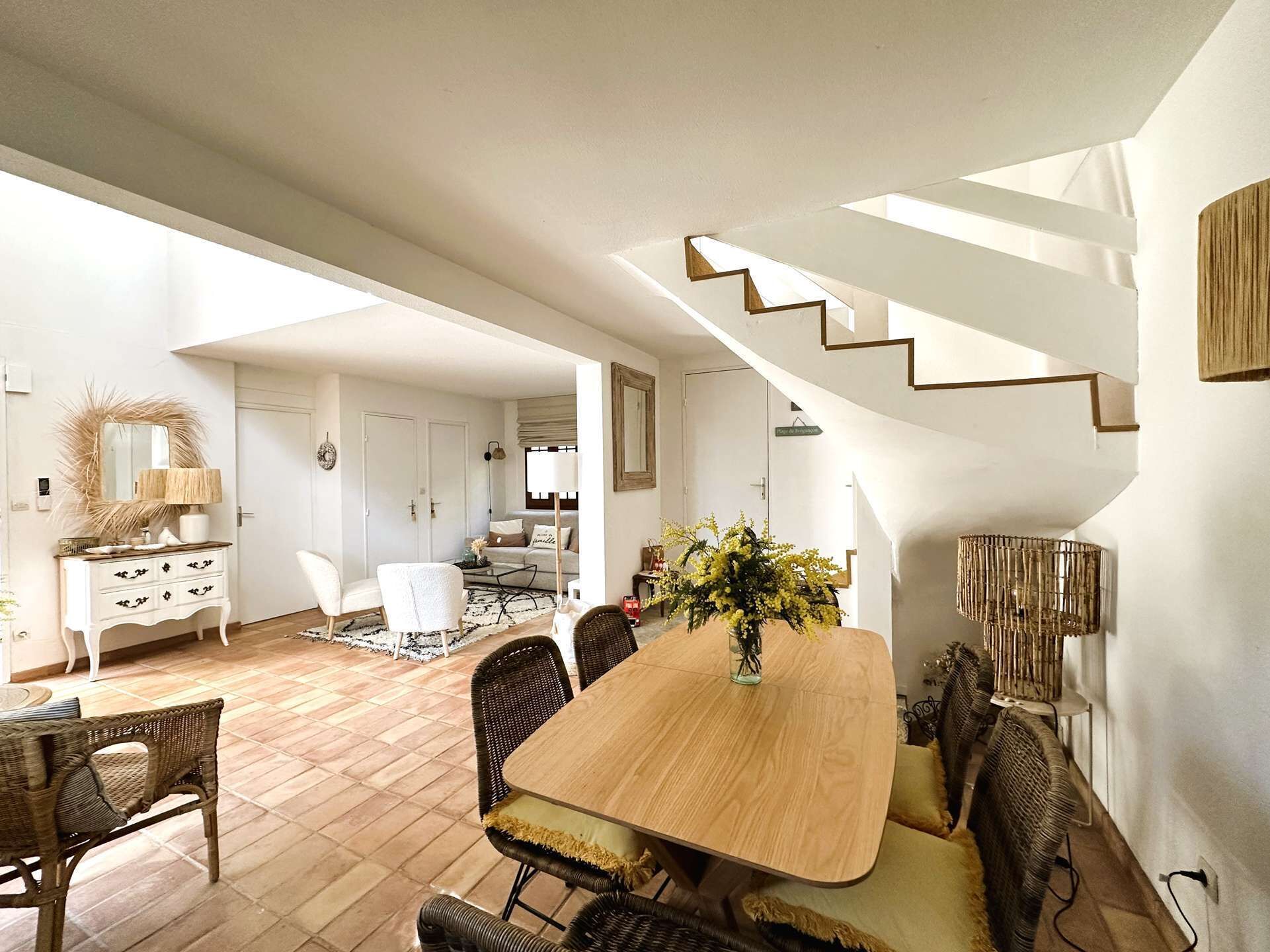 Un amplio salón con una mesa de comedor de madera, sillas de mimbre y una escalera que sube a un nivel superior.