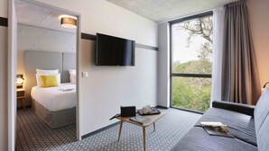Habitación de hotel moderna con una cama, televisión, una mesita con un libro, sofá y un ventanal con cortinas.