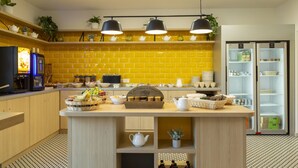Een moderne keuken met gele tegels, een centraal kookeiland met theeservies, een koffieapparaat en een koelkast.