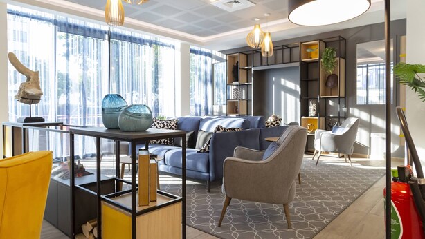 Een moderne woonkamer met een blauwe bank, een gele fauteuil en een glazen salontafel. Er zijn hanglampen, een boekenkast en een groot raam met gordijnen.