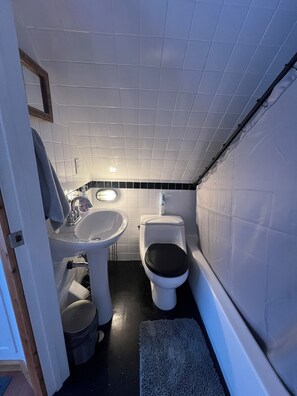 Een compacte badkamer met witte tegels, een wastafel, een toilet en een kleine prullenbak.