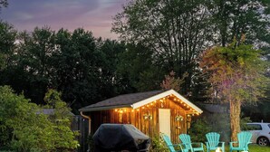 A wooden shed with string lights, a barbecue grill, and three turquoise chairs around a small fire pit in a garden. - Summer Beach Family Cottage  🏖 2min→beach @ Bamboo Chalet ~ Lake Erie 🇨🇦 
 (Turkey Point)