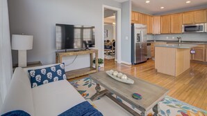 A modern living room with a sofa, a coffee table, and a television. The kitchen features wooden cabinets and stainless steel appliances.