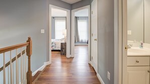 A hallway with wooden flooring, a staircase with white balusters, and a door leading to a bedroom with a bed and a nightstand.