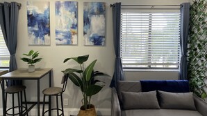 A modern living room with a sofa, a coffee table, and two bar stools. There are abstract paintings on the wall and a large window with blinds. - Sunny Palms Inn (Lake Worth)