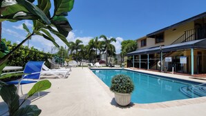 A swimming pool with lounge chairs and a covered seating area, surrounded by greenery and a building with a poolside bar. - Sunny Palms Inn (Lake Worth)