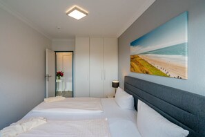 Ein Schlafzimmer mit zwei Betten, einem Nachttisch, einer Lampe, einem Strandbild und einer Tür zu einem anderen Zimmer.