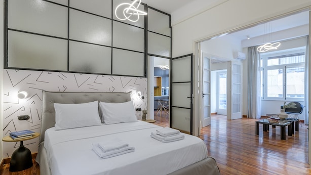 A modern hotel room with a large bed, a bedside table with books, and a view of the adjacent room. - 3Bdr Modern Design & Bright Interiors in Plaka (Athens)