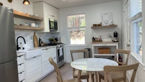 A modern kitchen with white cabinetry, a stainless steel refrigerator, a round marble table, and wooden chairs.
