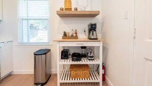 A compact kitchen with a coffee maker, a trash bin, and a shelf with a bottle and a basket.