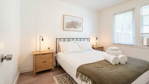 A bedroom with a bed, bedside tables, a nightstand, a window with blinds, and a framed picture on the wall.