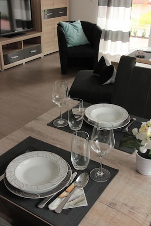Uma mesa de jantar posta com pratos, copos e talheres, um sofá e uma televisão numa sala de estar.