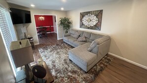 A living room with a grey sofa, a patterned rug, a flat-screen TV, and a dining area with a red wall and chandelier.