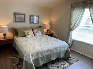 A bedroom with a bed, two nightstands, two lamps, two framed pictures on the wall, and a window with curtains.