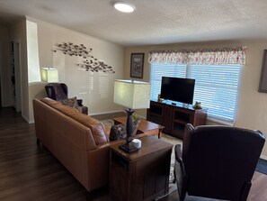 A living room with a brown leather sofa, a wooden coffee table, a TV on a stand, and a window with blinds.