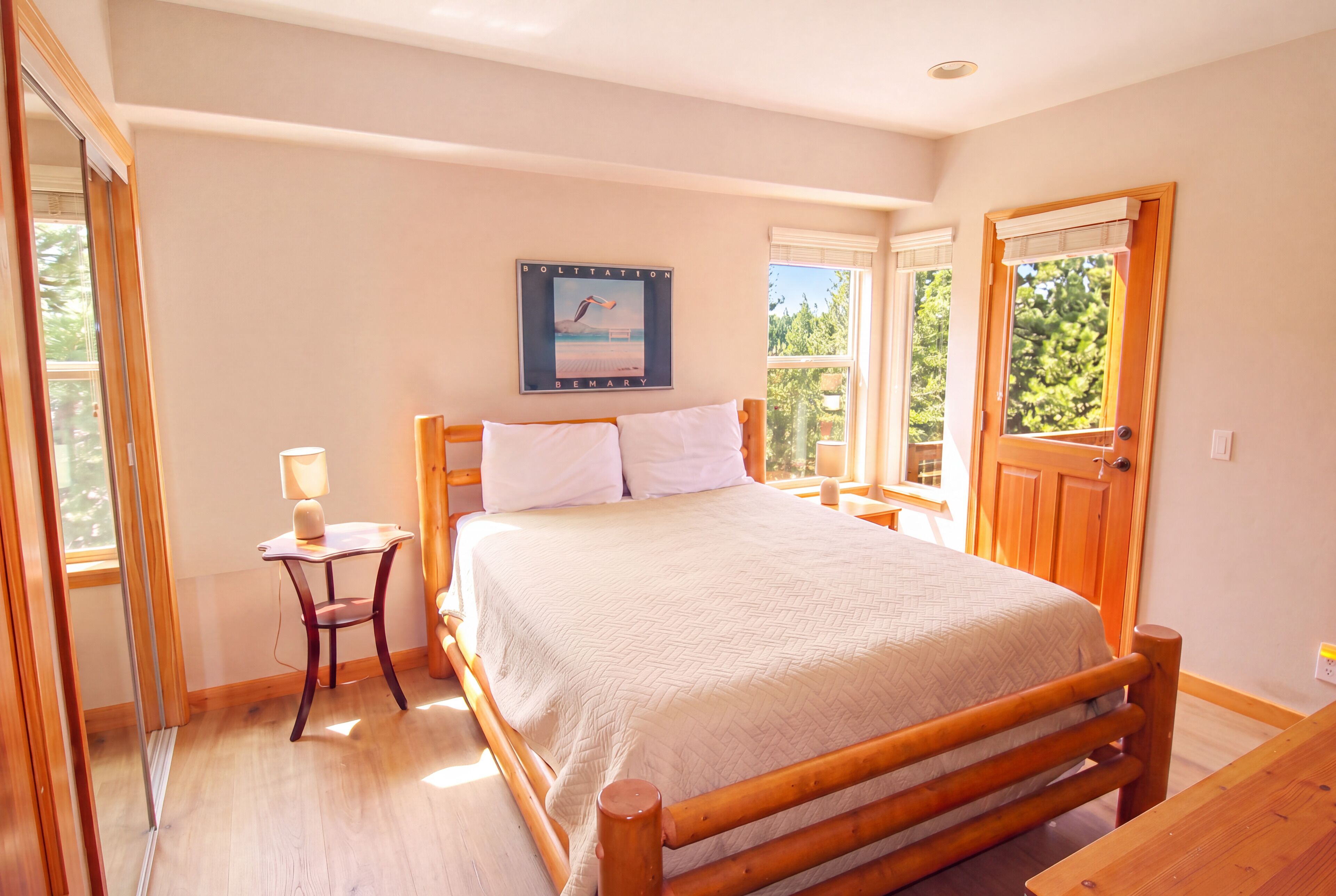 A bedroom with a wooden bed, a bedside table with a lamp, a window with a view of greenery, and a door.