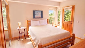 Un dormitorio con cama de madera, una mesita de noche con lámpara, una ventana con vista a follaje y una puerta.