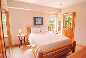 A bedroom with a wooden bed, a bedside table with a lamp, a window with a view of greenery, and a door. - Hideaway Down Canyon #110, End Unit close to June Mtn (June Lake)