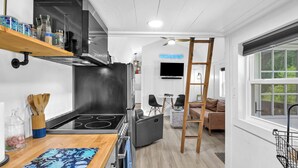 A compact kitchen with a stove, oven, and built-in microwave. There is a wooden shelf above the stove holding jars and utensils. A TV is mounted on the wall, and a ladder is propped against the wall.