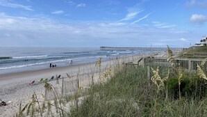 A beach with a pier extending into the ocean, sandy beach, and tall grasses in the foreground.