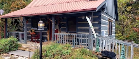 A rustic log cabin with a red metal roof, a stone chimney, and a porch with a railing.