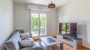 A modern living room with a grey sofa, a coffee table, and a TV on a wooden stand. There is a sliding glass door leading to a balcony with greenery outside. - Appartement Charly - Welkeys (Cabourg)