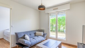 A modern, well-lit room with a grey sofa, a wooden coffee table, and a large window providing a view of greenery outside. - Appartement Charly - Welkeys (Cabourg)