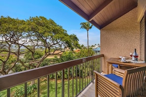 Un balcon donnant sur des arbres et des maisons, une table avec une bouteille et des verres, et une chaise.