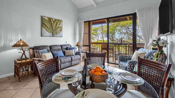 A spacious living area with a sofa, dining table, and chairs, a ceiling fan, and a large window with a view of trees. - Bright and Cheery Maui Vista 2 BR/2BA   MV2420 (Kihei)