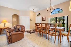 A spacious living room with a brown leather sofa, a dining area with wooden chairs and a table, and a large window with a view of palm trees.