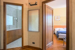 A hotel room with a wooden floor, a bathroom with a shower and sink, a bed with a blue blanket, and a wooden door.