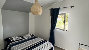 Ein Schlafzimmer mit Bett, einer Pendelleuchte, einem Fenster mit Vorhängen und Blick ins Grüne.