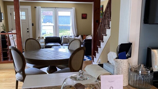 Un'area pranzo con un tavolo rotondo e sedie, un bancone cucina con una cupola in vetro che espone muffin e una scala che conduce a un altro piano.