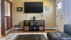 Un soggiorno con una grande televisione a schermo piatto montata a parete, un mobile TV nero con ripiani, un tappeto decorato e due poggiapiedi in pelle.
