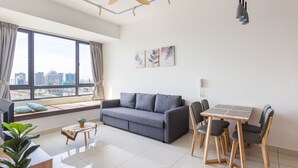 A modern living room with a grey sofa, wooden dining table, and chairs, large windows with a city view, and a ceiling fan. - Greenfield Residence at Bandar Sunway (Petaling Jaya)
