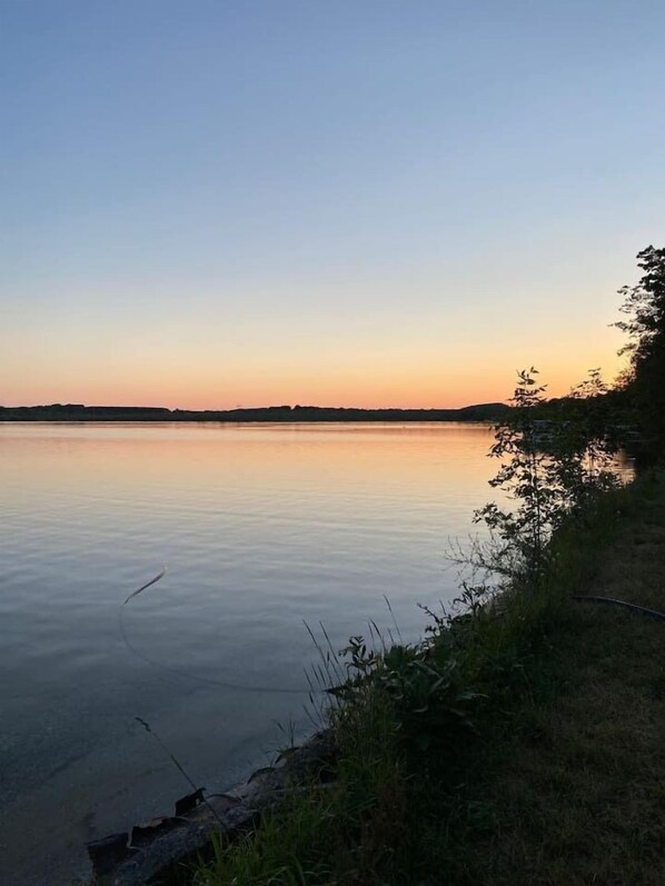 A calm lake at sunset with a grassy shoreline and distant hills.