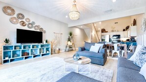 A modern living room with a flat-screen TV, blue shelving, grey sofas, and a kitchen in the background. - Salty Air, Ocean Views, Private Heated Pool - Steps to the Sand (Panama City Beach)
