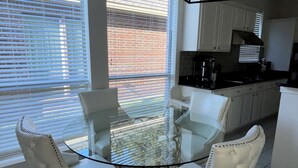 A dining area with a glass table surrounded by four white upholstered chairs with wooden legs.