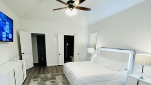 A bedroom with a bed, nightstands, a ceiling fan, a TV, and a door leading to another room.
